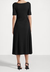 Svart knälång klänning med korta rullade ärmar och bred rund halsringning. Slätt tyg med en flytande silhuett. Höga, smala svarta klackar.