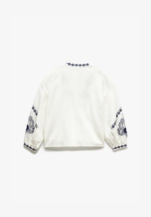 Valkoinen teksturoitu paita pitkillä puffi hihnoilla. Varustettu tummansinisin brodeeratuin yksityiskohdin hihansuissa ja kaula-aukossa. Neliömäinen leikkaus.