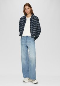 Niebiesko-czarny, wzorzysty krótki żakiet noszony na białej bluzce, w połączeniu z jasnoniebieskimi spodniami typu wide-leg i beżowo-szarymi sneakerami. Neutralne tło.