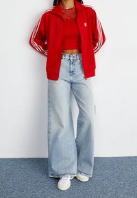 Czerwona bluza sportowa na zamek z białymi paskami, krótki czerwony top, jasnoniebieskie szerokie dżinsy, białe sneakersy oraz teksturowane detale.