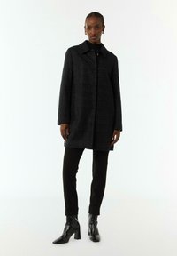 Black textured coat with a straight cut, large collar, and front closure. Paired with black fitted pants and pointed heeled boots.
