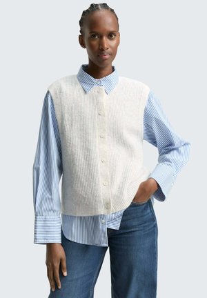 Un gilet en laine blanc tricoté par-dessus une chemise à boutons rayée bleue et blanche. Le gilet a une texture côtelée et des boutons devant. Un jean bleu complète le look.