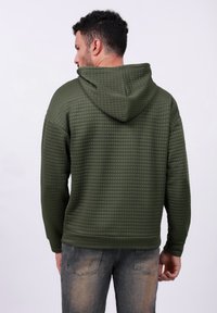 Sudadera acolchada verde con un patrón de cuadrícula texturizado, con capucha ajustable y puños acanalados, combinada con jeans azules deslavados.