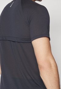 Detailní záběr na horní část zad a ruku osoby v tmavém, krátkém sportovním tričku s texturou, na kterém je u krku malé reflexní logo.