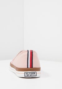 Zapatilla deportiva de lona rosa claro con suela blanca y detalles en marrón. Presenta un diseño a rayas en rojo, blanco y azul en el talón.