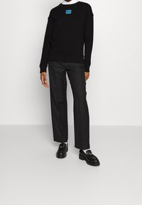 Persona che indossa un maglione nero con il logo blu "HUGO", pantaloni neri eleganti, calze bianche e mocassini neri con dettaglio oro, in piedi su un pavimento grigio.