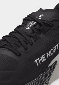 Κοντινό πλάνο ενός μαύρου αθλητικού παπουτσιού με υφασμένες κορδόνια, λευκή επωνυμία "THE NORTH" και τμήμα σόλας με σχέδιο.