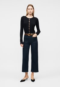 Γυναίκα που φοράει μακρυμάνικο μαύρο πουκάμισο με κουμπιά, σκούρα τζιν με φαρδιά πόδια, καφέ ζώνη και μαύρα παπούτσια με μυτερή μύτη και χαμηλό τακούνι, στέκεται με το ένα χέρι στην τσέπη.