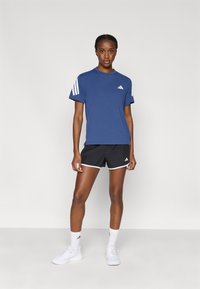 Μπλε αθλητική μπλούζα με λευκές ρίγες, συνδυασμένη με μαύρο σορτς με λευκή επένδυση. Το μοντέλο φοράει λευκά αθλητικά παπούτσια και κάλτσες.