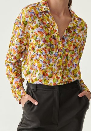 Kvinde iført en farverig, abstrakt blomstret skjorte med knapper, stoppet ned i sorte læderbukser, med hænderne i lommerne.