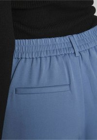 Gros plan sur un pantalon bleu avec ceinture élastique et poche passepoilée, porté avec un haut noir côtelé.