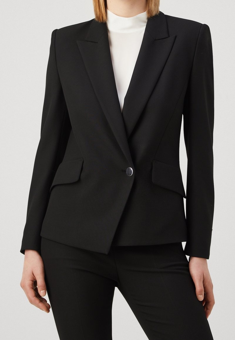 Černý oblekový blazer s ostrými klopami, zapínání na jeden knoflík, dvě přední kapsy a strukturované ramena. Nosený přes bílou halenku.