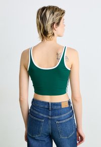 Grön crop top med vita detaljer, tillverkad i bomullsblandning. Bärs med högmidjade blå denimjeans. Synlig tatuering på ryggen.