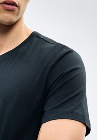 Κοντινό πλάνο ενός ατόμου που φοράει απλό μαύρο t-shirt με κοντά μανίκια, δείχνοντας τον ώμο, το πάνω μέρος του στήθους και μέρος του προσώπου.