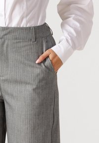 Persoon met grijze pantalon met fijne strepen en een wit overhemd met knopen, hand rustend in de zak van de broek, op een effen achtergrond.