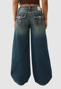 Jeansy o szerokich nogawkach w ciemnym odcieniu niebieskiego denimu z przetarciami, zdobione dekoracyjnym przeszyciem oraz dwoma tylnymi kieszeniami z wzorzystymi akcentami.