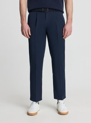 Mężczyzna ubrany w granatowe, dopasowane spodnie, czarny pasek oraz białe trampki z brązowymi podeszwami, stojący na jednolitym jasnym tle.