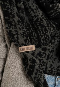 Zwarte en grijze gebreide sjaal met een zachte textuur. Een beige label met de tekst "Knit Factory." De achtergrond heeft een textuur van beige stof.