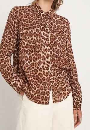 Vrouw die een lang mouw, knoopjes-horend overhemd met luipaardprint draagt, ingestopt in een witte broek, met haar hand in de zak en een ring aan haar vinger.