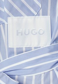 Jasnoniebieska tkanina z białymi paskami, z białą haftowaną etykietą z napisem "HUGO". Zawiera pasujący materiałowy pasek.