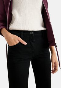Jeans neri a taglio slim con dettagli di cucitura sottili, abbinati a un maglione grigio chiaro a coste e una giacca viola.