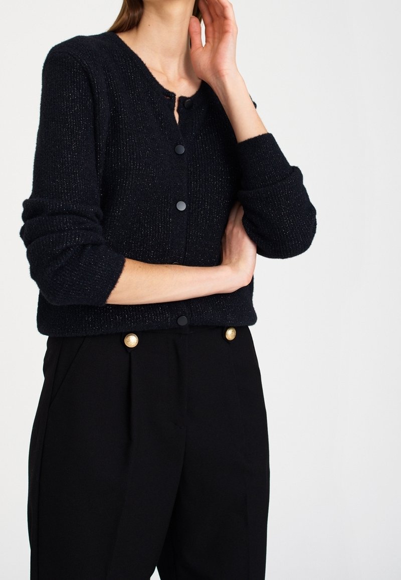 Marinblå stickad cardigan med rund halsringning, långa ärmar och svarta knappar. Bärs över svarta högmidjade byxor med guldaccenter.