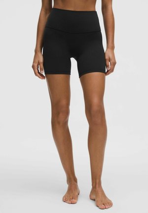 Kobieta w wysokich czarnych krótkich spodenkach rowerowych stojąca boso na jednolitym jasnoszarym tle.