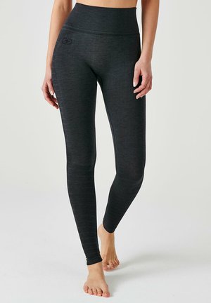 Vrouw draagt hooggetailleerde, donkergrijze naadloze legging en staat blootsvoets tegen een eenvoudige lichte achtergrond.