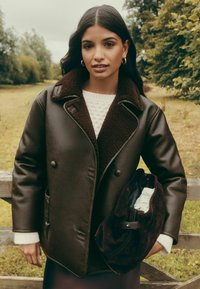 Pruun nahast jakk tumedapruuni lambanaha kraega, kahekordsete nööpidega ja küljetaskutega. Modell kannab tumedat seemisnahast kotti.