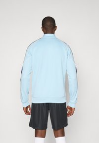 Light blue athletic jacket with a high collar and black accents on the shoulders. The fabric appears smooth, complemented by elastic cuffs.