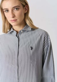 Camicia a righe grigie e bianche con bottoni e colletto, con un piccolo logo nero ricamato sul petto. Texture liscia.