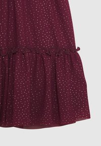 Bordeaux stof met kleine gouden stippen en een ruchesafwerking bij de zoom op een semi-doorzichtige textuur.