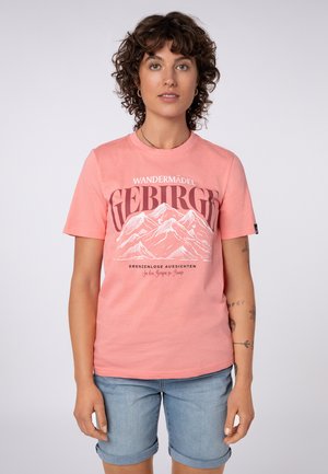 Frau mit kurzen lockigen Haaren, die ein pinkes T-Shirt mit Bergmotiv und hellblaue hochgekrempelte Jeansshorts trägt, steht vor einem schlichten Hintergrund.