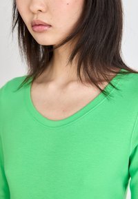 Haut long en coton vert avec un col rond, présentant une texture lisse et une coupe ajustée. Gros plan sur le col et la zone des épaules.