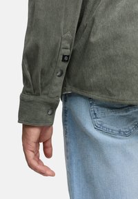 Camisa de pana verde con un corte entallado, con puños abotonados, etiqueta negra y superficie texturizada, combinada con vaqueros de mezclilla azul claro.