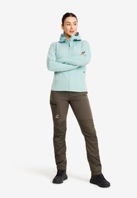 Kvinna står med armarna i kors, iförd en ljusblå zip-up hoodie, bruna cargo-byxor och svarta vandringsskor på en vit bakgrund.