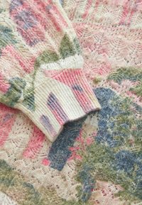 Gehaakt trui fragment met bloemmotieven in pasteltinten op een gestructureerde achtergrond. Ribbelmanchet in roze en crème.