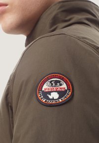 Oliven grøn jakke med en tekstureret overflade, der har et rundt broderet mærke, som siger "Antarctic Research Program" i rød og orange.