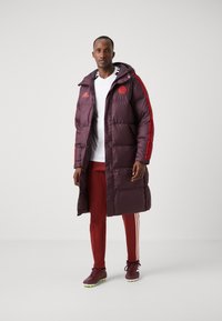 Abrigo morado esponjoso con rayas rojas, capucha y logo, combinado con pantalones deportivos rojos y botas de fútbol marrones. Camiseta blanca simple debajo.