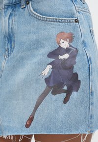 Το τζιν φούστα διαθέτει μπλε πλύση, ξεφτισμένο τελείωμα και τυπωμένο σχέδιο ενός χαρακτήρα με σκούρο ντύσιμο, που κρατάει όπλο, σε πόζα δράσης.