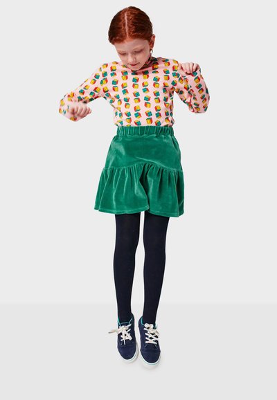 Chemise à manches longues avec un motif en blocs colorés, associée à une jupe en velours vert à volants. Portée avec des collants noirs et des baskets bleu marine.