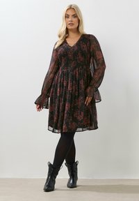 Musta kukkamekko, jossa on läpikuultavat pitkät hihat, rypytetty yläosa ja kerroksellinen hameosa. Yksityiskohtina punaisia kukkakuvioita ja vaaleat hihansuut.