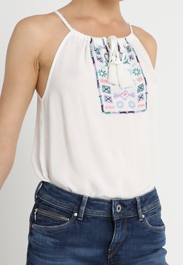 Biała bezrękawnik z gromadzonym dekoltem, zdobiona kolorowym haftem geometrycznym i frędzlami na środku. Połączona z dżinsowymi szortami.