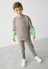 Grauer Sweatshirt- und Hosenanzug mit mintgrünen Akzenten, großen "BAKER"-Schriftzug, weicher Textur, Reißverschlusstasche und weißen Sneakers.