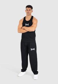 Schwarzes ärmelloses Tanktop und schwarze Jogginghosen mit weißem "SMILODOX"-Logo. Das Model trägt weiße Sneakers und steht mit verschränkten Armen.