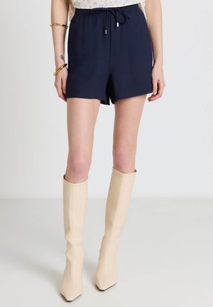 Donna che indossa pantaloncini con coulisse blu navy, stivali crema al ginocchio, braccialetti d'oro e una maglietta floreale tagliata sopra la vita.