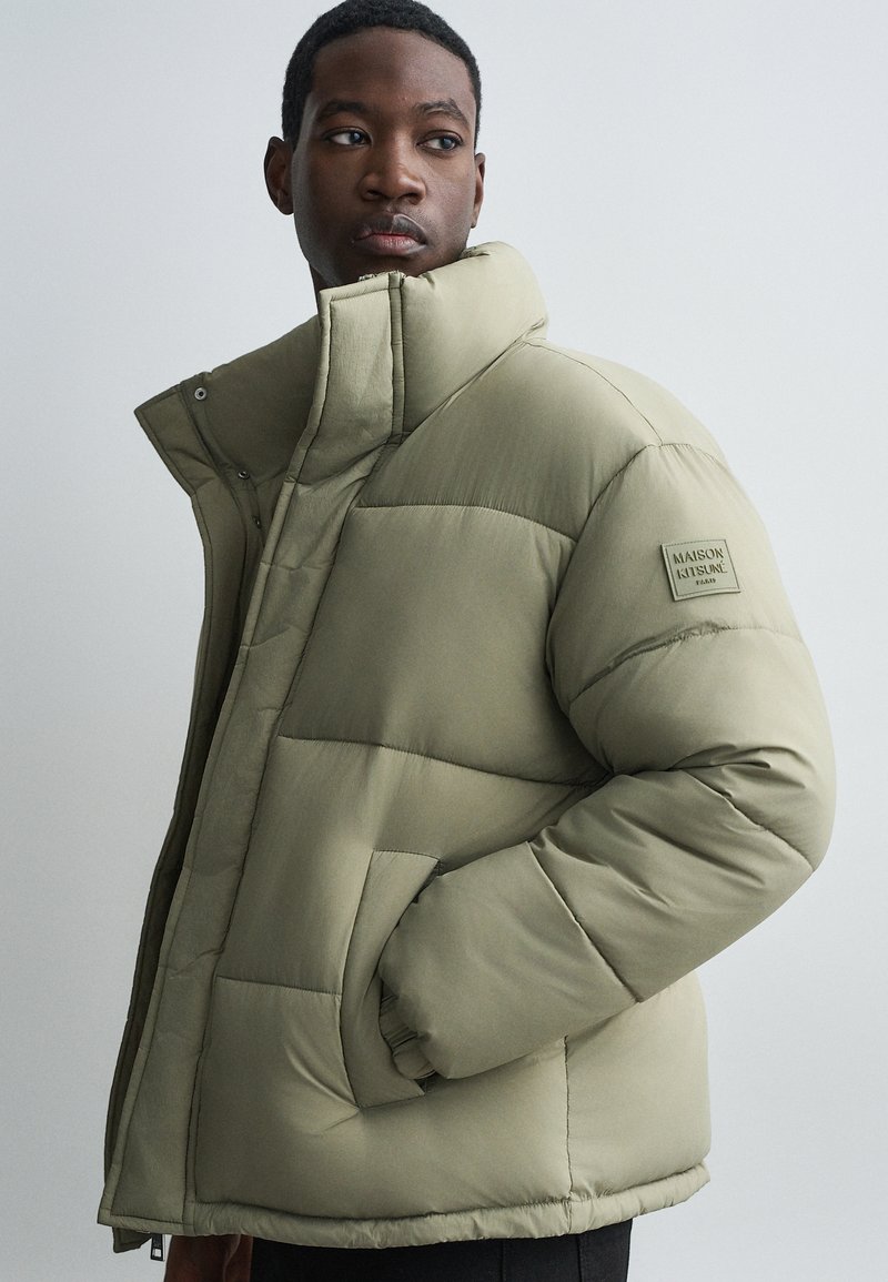 Oliwkowa puchowa kurtka z wysokim kołnierzem, pikowanym wzorem, bocznymi kieszeniami i naszywką z logo na lewej rękawie. Teksturowana faktura materiału.