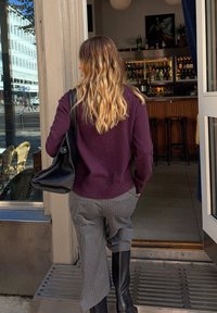 Purpurinė megztinis, pilkos dryžuotos kelnės iki kulkšnies ir juodos kelio ilgio botai. Juoda rankinė per petį; matomas baro įėjimas.