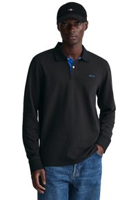 Schwarzes Langarm-Polohemd mit blauem Kragen und GANT-Logo; kombiniert mit hellblauen Jeans; Modell trägt eine schwarze Mütze.