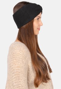 Bandeau en polaire noire avec un design en nœud torsadé, texture douce et bande large. Modèle le portant avec des cheveux longs et raides et un pull léger.